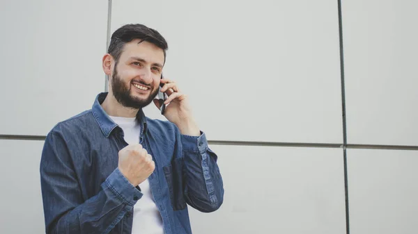 Happy young bearded man talking on mobile phone and delighted clench fist victory triumph gesture, hearing good news in conversation standing on grey wall background. Celebrate success, achieve goal.