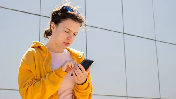 Serious confused young woman in yellow hoodie holding mobile phone and staring at screen,standing outdoors, having problem with internet, reading bad news in message, annoyed by missed call or spam.