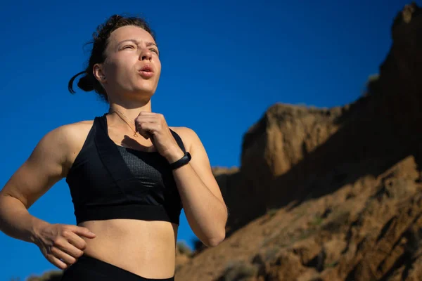 Siyah spor giyim maratonunda koşan genç bir kadın inanılmaz dağ manzarası ve mavi gökyüzünde derin bir nefes alıyor. Triatlon koşusu ve aktif yaşam tarzı eğitimi. Boşluğu kopyala.