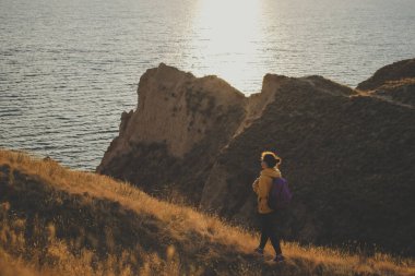 Sırt çantalı, sarı kapüşonlu genç bir gezgin dağ zirvesinde patikada yürüyor, gün batımında deniz kenarındaki uçuruma yürüyor, doğa manzarasının tadını çıkarıyor. Yumuşak filtre. Macera konsepti