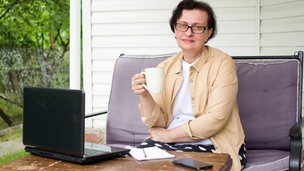 집 테라스에 있는 소파에 앉아 컴퓨터를 쓰고 카메라를 보며 쉬고 있는 중에 안경을 끼고 커피를 마시고 있는 노인의 모습. 연로 한 프리랜서 노동자 개념 — 스톡 사진