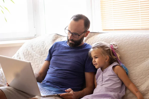 Young bearded father with small kid daughter looking at laptop screen, watching cartoons, educational video, shopping online using wireless internet connection while sitting on sofa at home