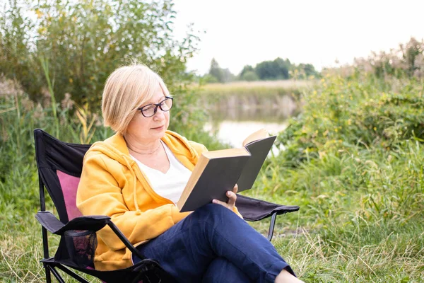 Mooie blonde senior vrouw in bril en gele hoodie lezen interessant boek bestseller zitten in toeristische stoel op de camping tijdens het weekend natuur picknick in de buurt van meer of rivier buiten — Stockfoto