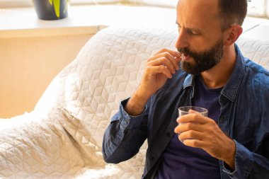 Milenyum sakallı bir adam bir bardak suyla ağrı kesiciyi dindirmek için antibiyotik alıyor, hasta hissediyor, evinde kanepede otururken baş ağrısı çekiyor. Acil tedavi kavramı