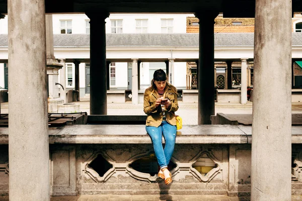 Hipster young woman sitting at the old fish market of an historic european town holding a mobile phone and typing a message