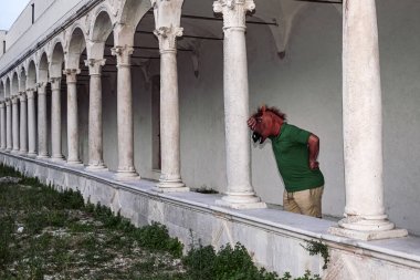 man with a horse head mask is leaning against an column under an ancient cloister while thinks about his life. Problems, personality, mysticism, thoughts, life concept clipart