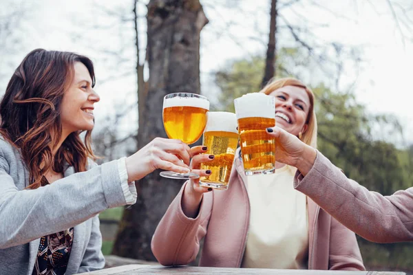 three women sitting at an outdoor terrace in autumn, talking, laughing and toasting together with beer.