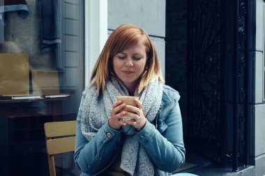 Genç, kızıl saçlı bir kadın dışarıda oturmuş gülümsüyor ve ellerinde lezzetli bir fincan kremalı kahve tutuyor.