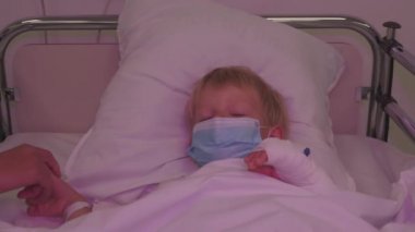 Acil bir durumda hastane yatağında yatan küçük bir çocuk. Ambulans ışıklarıyla aydınlatılmış. Annesi hastanede maske takan hasta bir çocuk.