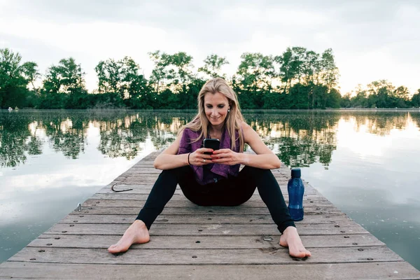 cheerful athletic woman relaxing after workout - smiling woman using mobile phone after sport - caucasian woman in sportswear sitting on the pier