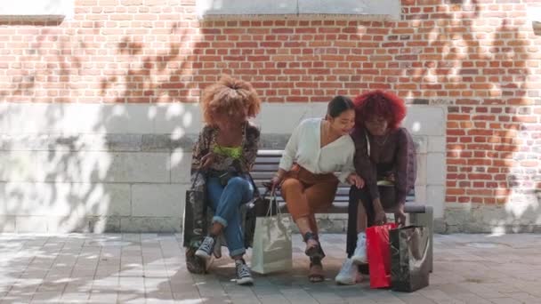 市内中心部のベンチの屋外に座っている多人種の女の子買い物袋を楽しみながら話しています ライフスタイル友情買い物ゲンツのコンセプト — ストック動画
