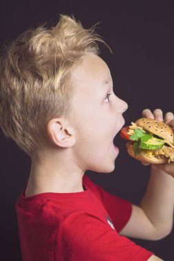 Kapalı ağızlı komik çocuk tavuk, sebze ve peynirli lezzetli bir burger yiyor.