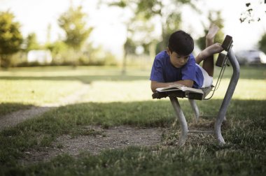 Giovane ragazzino sudamericano mulatto legge un libro sdraiato su una panchina nel parco sotto casa