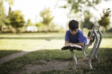 Giovane ragazzino sudamericano mulatto legge un libro sdraiato su una panchina nel parco sotto casa