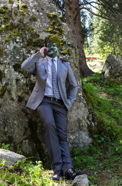 Un impiegato in giacca e cravatta, all'aperto con un trucco in faccia e muschio verde in testa. L'uomo si fonde con la natura . — Foto Stock