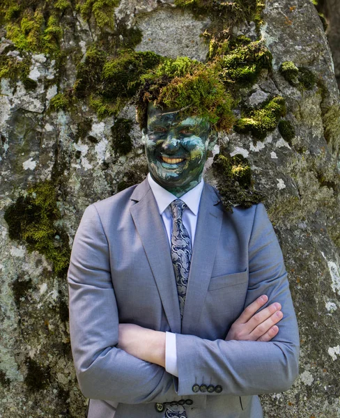 Office worker in a business suit, outdoors with a make-up on his face and green moss on his head. Man merges with nature.