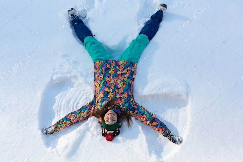 High angle view of happy girl lying on snow and moving her arms and legs up and down creating a snow angel figure. Smiling woman lying on snow in winter holiday with copy space