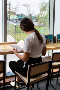 Şık gözlük ile şık coworking uzayda açık kitap masada otururken hayal olumlu genç kadın. Uzak bestseller okuma sırasında seyir gözlük güzel kadın gülümseyerek