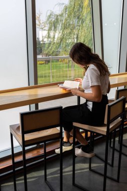 Şık gözlük ile şık coworking uzayda açık kitap masada otururken hayal olumlu genç kadın. Uzak bestseller okuma sırasında seyir gözlük güzel kadın gülümseyerek