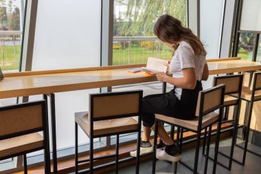 Şık gözlük ile şık coworking uzayda açık kitap masada otururken hayal olumlu genç kadın. Uzak bestseller okuma sırasında seyir gözlük güzel kadın gülümseyerek