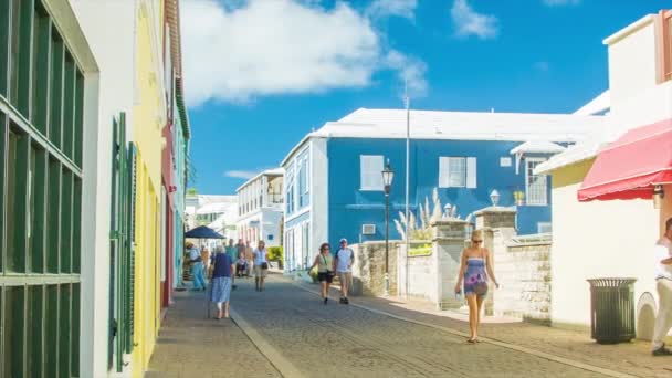 歴史的な町が牧歌的な日に提供しているものを探索するカラフルなバミューダンの建物の間の石畳の道を歩く人々とセントジョージの町バミューダの活気あるサイドストリートシーン — ストック動画