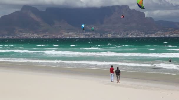 南アフリカのケープタウンのブルーベルクビーチに立つ二人の男性が テーブルマウンテンとケープタウンの前の街の景色を楽しんでいます 海の極端なカイトサーファーが見られます — ストック動画
