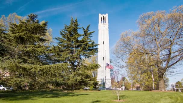 ノースカロライナ州ローリーにあるNc州立大学の記念ベルタワーは 晴れた青空の背景の日に木々とアメリカ国旗に囲まれています — ストック動画