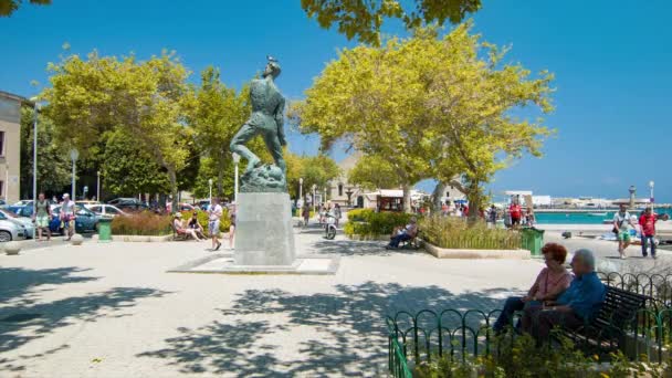 歴史的な彫像を持つ公園の設定を散策する晴れた日に活気に満ちたマンドラキ港エリアを楽しむロードスギリシャの人々 — ストック動画