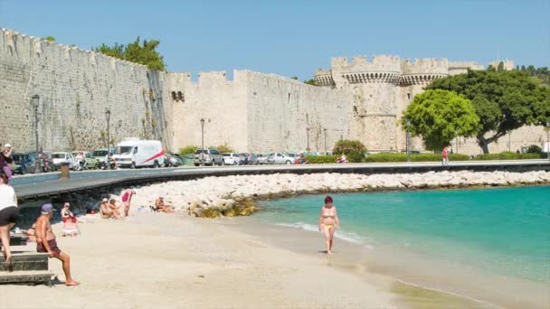 古代旧市街の城壁の背景と観光客が晴れた日に青いエーゲ海の水に沿って歩いて楽しむアクティサクトゥーリのロードスギリシャビーチクローズアップ — ストック動画