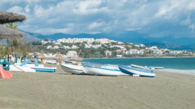 Arka Planda Apartment Resort Binalar ve Dağlar ile Güneşli Bir Günde Kum üzerinde Eğlence Tekneler ve Çim Kulübeler ile Estepona yakınlarındaki Güney İspanya Vacation Beach Scene