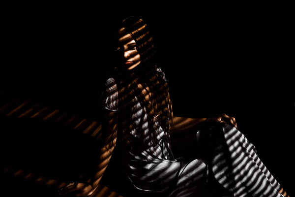 A beautiful girl in a dark room, light falls on her through the blinds, forming a striped pattern on the silhouette.
