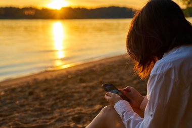 Gün batımında kumlu kıyı üzerinde oturan ve onun Smartphone ekranına bakarak Genç kız. Yalnız seyahat eden kadın onu cep telefonu kullanarak 
