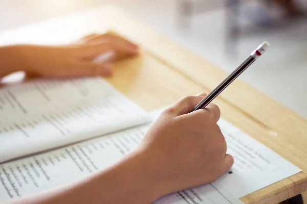 Students in uniform are doing the test in the classroom. Close up hand holding a pencil to write the answer to the exam on wooden table. Evaluation of learning for educational development