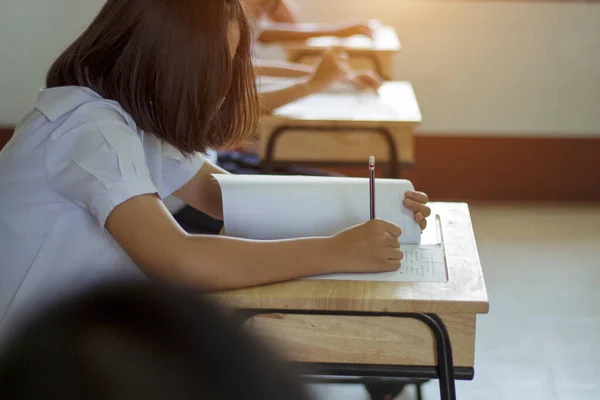 Students in uniform are doing the test in the classroom. Close up hand holding a pencil to write the answer to the exam on wooden table. Evaluation of learning for educational development