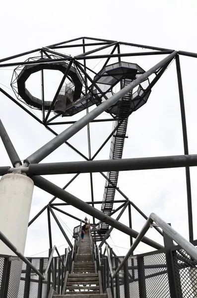 Passos que levam ao topo do Tetraedro em Bottrop, Alemanha, tomados com pessoas subindo escadas e céu branco no fundo. A estrutura de aço walkable é um marco local popular — Fotografia de Stock