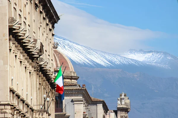 Historical building in Catania, Sicily, Italy with waving Italian flag. In the background cupola of famous Saint Agatha Cathedral and Mount Etna with snow on the very top of the volcano
