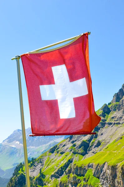 Vertical picture of Swiss waving flag with summer mountain landscape in background. National symbol. Swiss Alps. White cross on red field. National concept. Switzerland summer