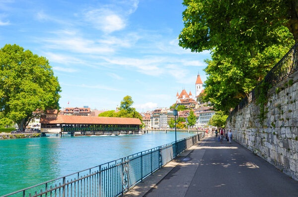 Thun, Switzerland - August 8, 2019: People walking on a riverside promenade in the Swiss city. Located by Bern, not far from the Swiss Alps. The dominant is a famous castle tower. Aare river.