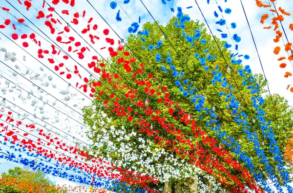 Schöne bunte Straßendekoration in loreto, madeira, portugal. Feier des religiösen Festes. rote, weiße, blaue und orangefarbene Papierblumen hängen in der Luft. Grüner Baum im Hintergrund — Stockfoto