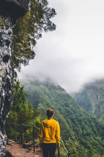 Jovem em camisola amarela no miradouro da Levada Caldeirao Verde, Madeira, Portugal. Montanhas panorâmicas verdes em névoa, paisagem nebulosa. Viajante feminino. Instagram, filtro hipster. Foto vertical Imagem De Stock