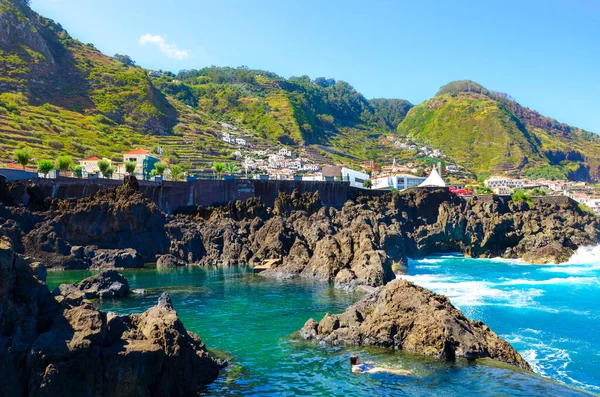 Vrouw die zwemt in natuurlijke zwembaden in de Atlantische Oceaan, Madeira, Portugal. Gemaakt van vulkanisch gesteente, waarin de zee van nature stroomt. Village Porto Moniz op de rotsachtige heuvel op de achtergrond — Stockfoto