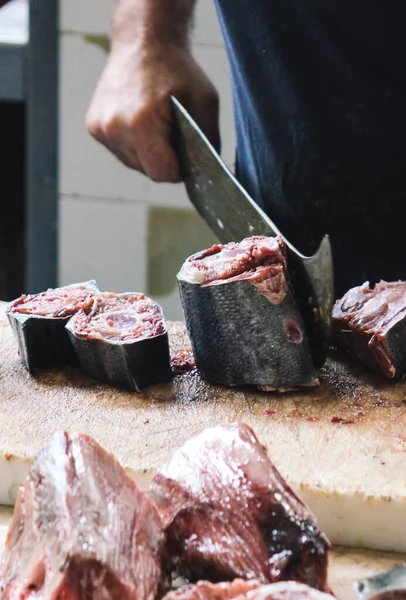 Detalle de un pescador cortando atún con una enorme cuchilla en una tabla de madera. Carne de atún desenfocada en primer plano. Mercado de pescado tradicional en Funchal, Madeira, Portugal. Industria pesquera, pesca —  Fotos de Stock