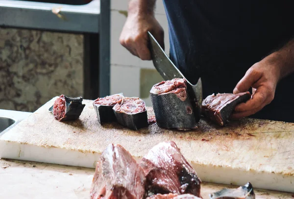 Cierren las manos del pescador cortando el atún con la cuchilla enorme sobre la tabla de madera. Carne de atún desenfocada en primer plano. Mercado local de pescado en Funchal, Madeira, Portugal. Derechos de los animales, abuso, crueldad —  Fotos de Stock
