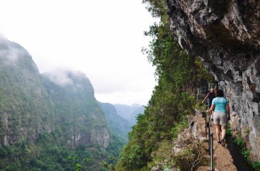 Levada do Caldeirao Verde Patikası sırasında kayanın kenarında dar bir patikada yürüyüş yapan turistler. Yeşil tropikal ormanlarla kaplı kayalar ve dağlar. Tehlikeli bir yürüyüş, uzun bir yürüyüş. Etkin tatil