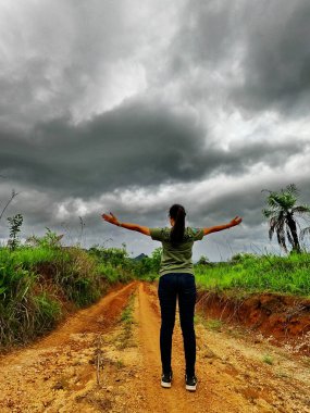 Genç kadın toprak yolun ortasında duruyor ve ellerini kaldırıyor. Güzel, sağlıklı, pofuduk saçları olan mutlu bir genç kız.