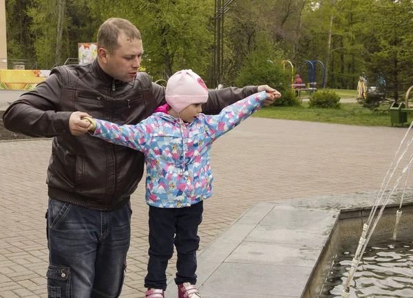 girl walks on the street. A girl and a dad are standing near the fountain. The girl reaches for the water in the fountain.