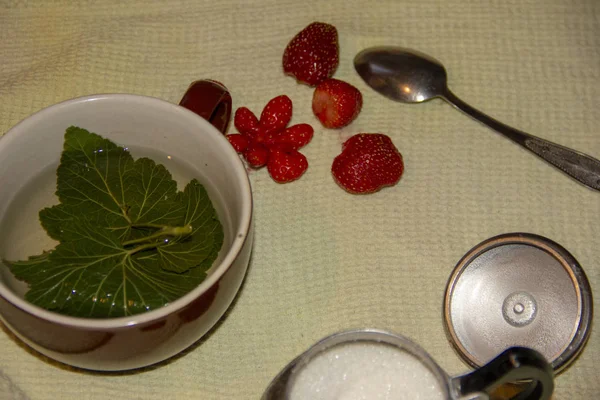 En la mesa hay una taza, un tazón de azúcar, una cuchara y una fresa. En una taza se encuentra un trozo de grosella y té vertido. hora del té . — Foto de Stock