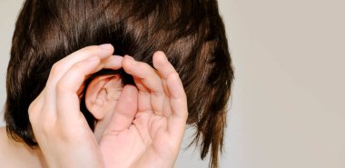 Dedikodu, söylenti, fısıltı, duyma konsepti. Genç kadın daha iyi duymak için elini kulağına sokuyor. Stüdyo çekimi gri arka planda. Yakın plan, kopyalama alanı