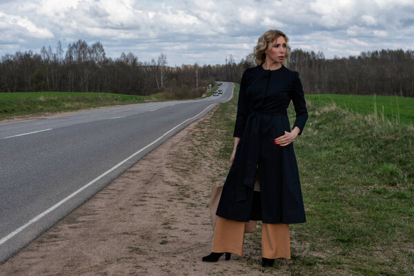 A middle-aged Caucasian woman in an elegant summer black coat stands alone near a highway in the countryside against a cloudy sky on a spring day. Copy space for your text or design.