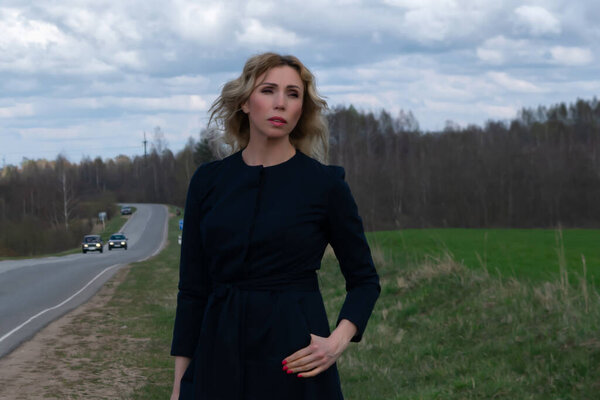 A middle-aged Caucasian woman in an elegant summer black coat stands alone near a highway in the countryside against a cloudy sky on a spring day. Copy space for your text or design.